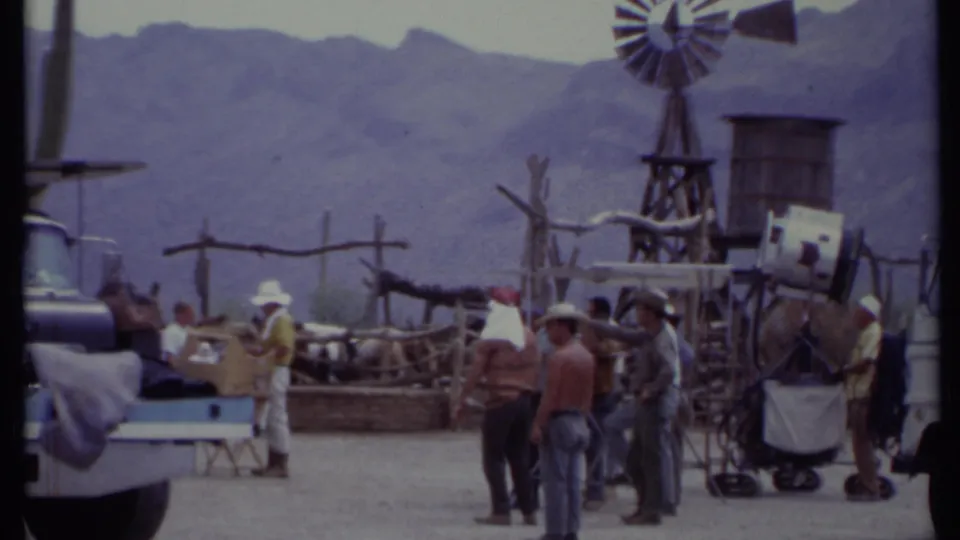 1971 California frame with several crew members in hats near equipment, a rustic set, windmill, water tank, and distant mountains.