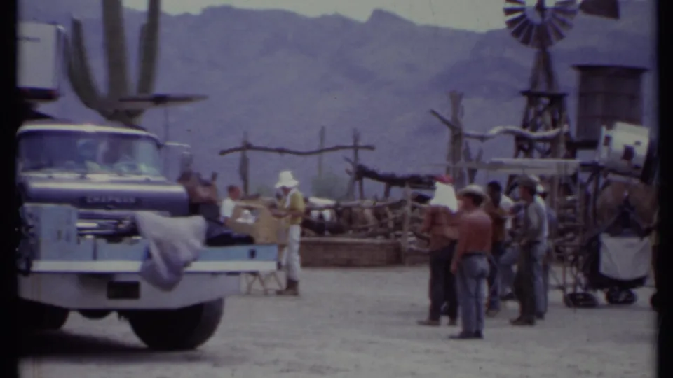 1971 California frame of crew members around a Chevrolet truck, rustic Western fencing, cactus props, windmill, and mountains.