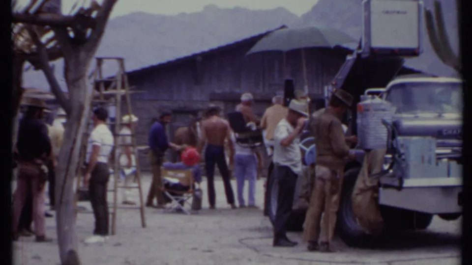 1971 California 8mm frame showing crew, ladders, chairs, a truck, and Western set structures in a desert production area.