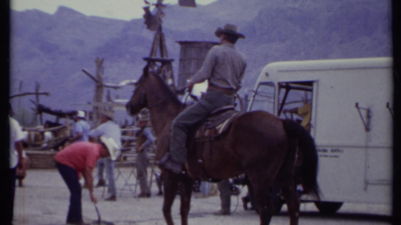 Cowboy-hatted rider on a brown horse near tourists and a white vehicle in a mountain setting.