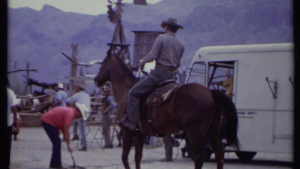 1971 California 8mm frame of a cowboy-hatted rider on a brown horse beside tourists, a white vehicle, and rustic structures.