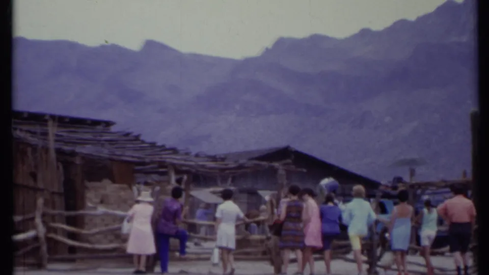 1971 California 8mm frame showing a group of tourists moving through a rustic Western-style attraction beneath rugged mountains.