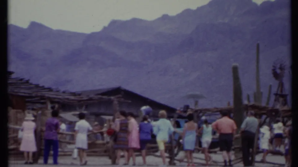 1971 California 8mm frame of tourists gathered along a rustic fence with cacti, a windmill, wooden structures, and mountains visible.