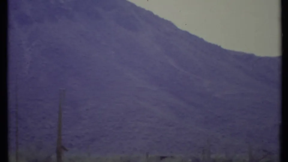 1971 8mm desert view in California showing a purple mountain range, pale sky, and sparse cactus forms in the foreground.