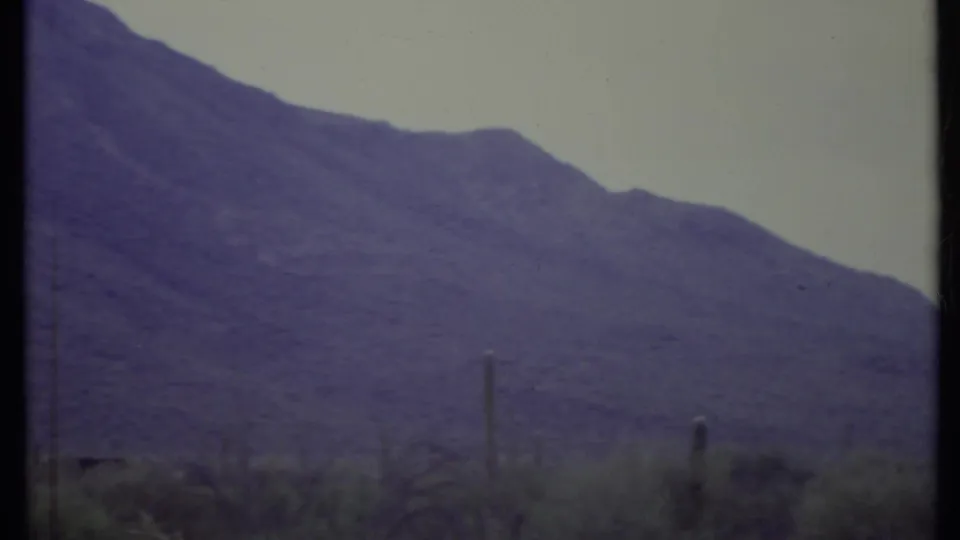 California desert landscape from a 1971 8mm home movie, with mountains, pale sky, scrub, and cactus silhouettes.
