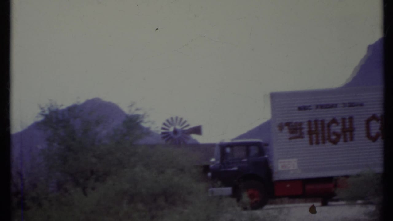 Dark semi truck parked near a windmill with desert bushes and mountains behind it.