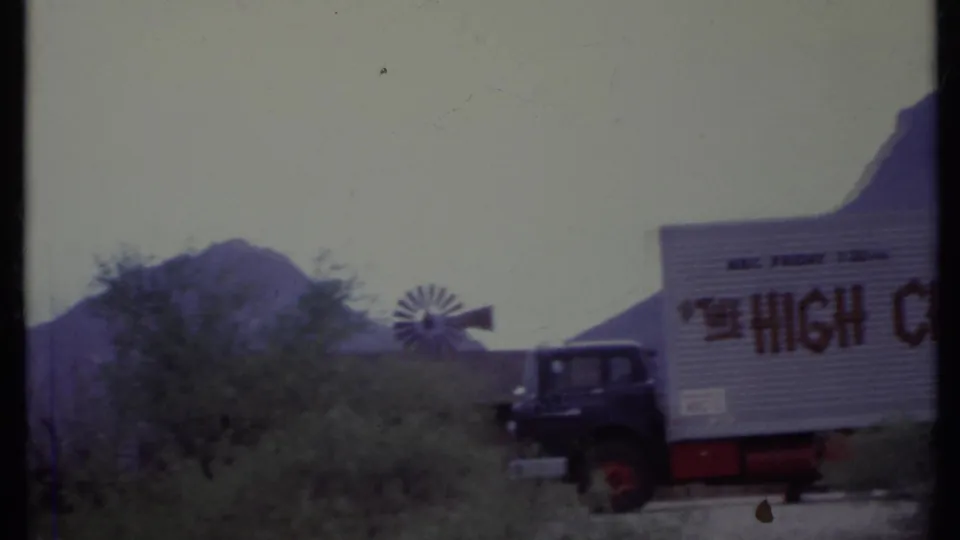 1971 California 8mm frame showing a parked dark semi truck with partial lettering, a small windmill, scrub vegetation, and hazy mountains.