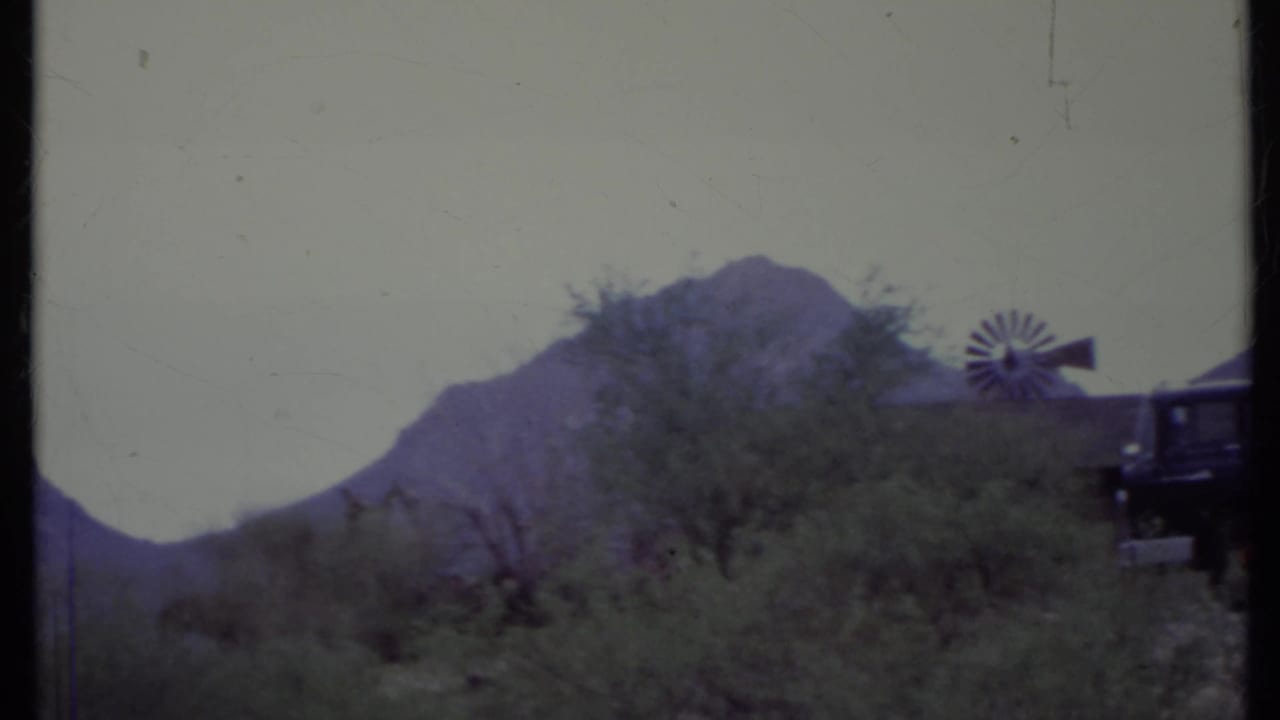 1971 California desert frame with a small windmill, scrub trees, rugged mountains, and the rear edge of a dark truck entering the frame.