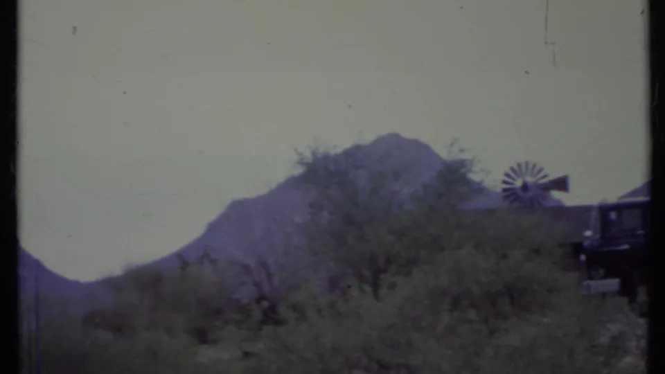1971 California desert frame with a small windmill, scrub trees, rugged mountains, and the rear edge of a dark truck entering the frame.