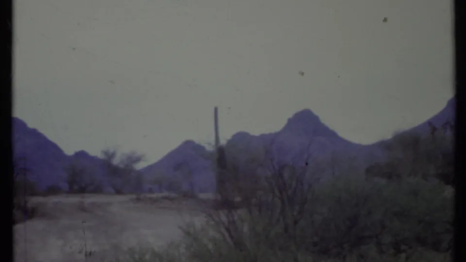 1971 California desert frame showing a pale road, low bushes, utility or fence posts, and distant rugged mountains in soft 8mm color.