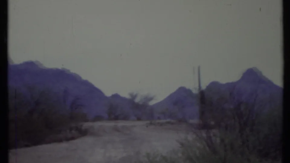 1971 California 8mm frame showing a pale dirt road, sparse desert vegetation, utility poles, and mountains under a washed sky.