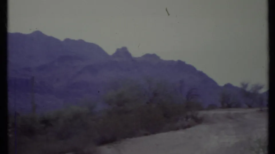 1971 California desert view with a road edge, scrub vegetation, cactus-like plants, and a large mountain ridge in soft focus.