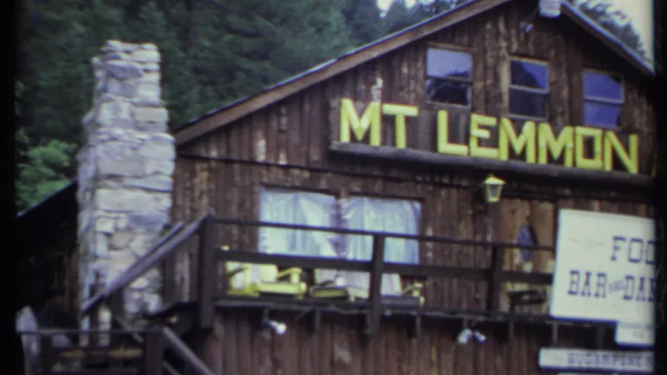 Rustic Mt Lemmon Inn exterior with yellow lettering, stone chimney, balcony rails, and surrounding trees in 1971 8mm footage.