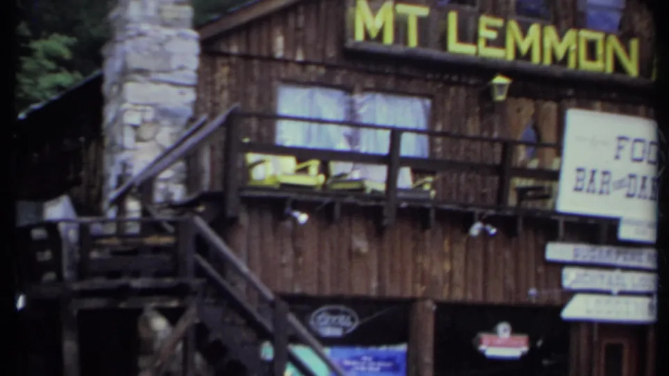 The camera frames the wooden inn exterior, stone chimney, balcony, stairs, Mt Lemmon lettering, and signs for food, lounge, lodging, and rooms.