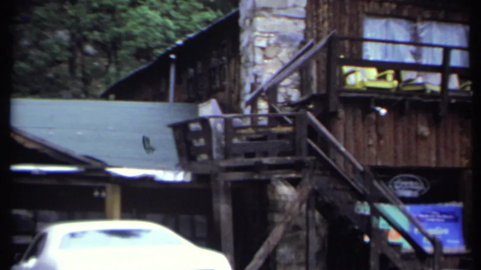 1971 home-movie frame shows the inn balcony, exterior stairs, stone chimney, wooded backdrop, and a white car passing or parked in front.