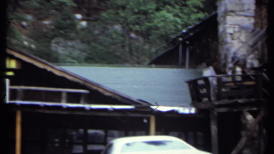 1971 8mm view of a rustic mountain inn exterior in California, with timber buildings, trees, and a white car near the frontage.
