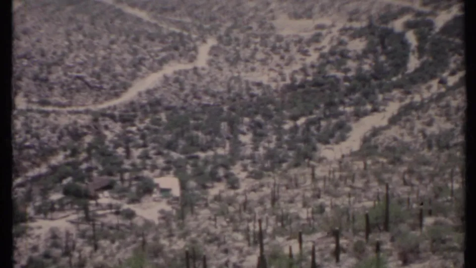 1971 8mm mountain landscape showing winding pale roads, scrub-covered slopes, tall cactus-like plants, and a small light structure below.
