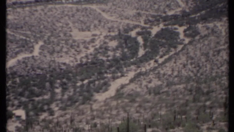 1971 8mm wide view of arid mountain terrain with intersecting dirt roads, scrub growth, and a small pale structure at lower left.
