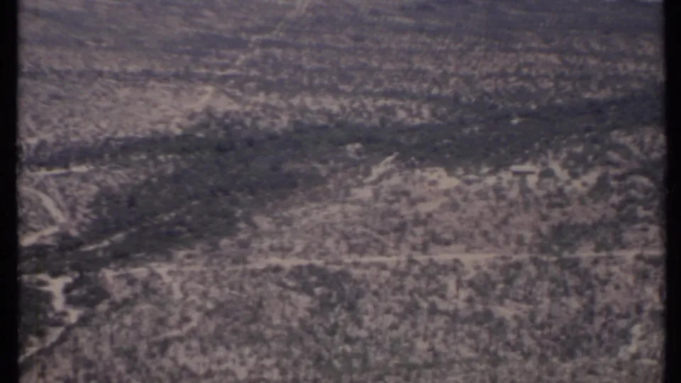 1971 California aerial footage frame with scattered scrub, pale road lines, and muted 8mm film texture.