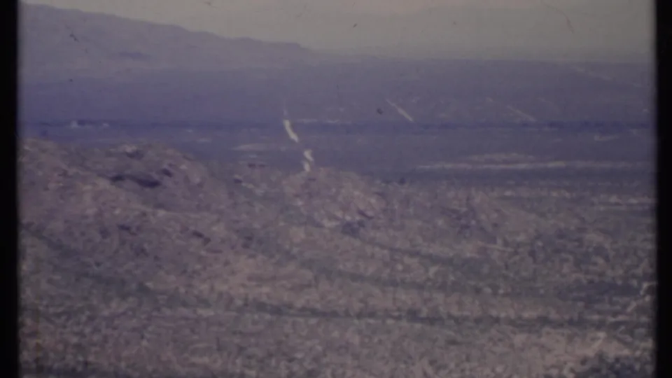 1971 8mm California desert view with a rocky foreground slope, sparse shrubs, pale tracks, and hazy mountains in the distance.