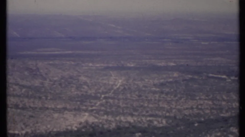8mm-era California desert frame with a rocky lower slope, open plain, faint road or wash lines, and hazy mountains beyond.