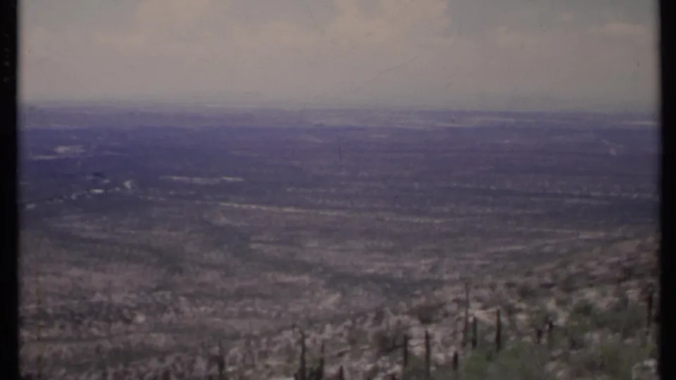 1971 California 8mm frame showing a hazy arid valley with sparse shrubs and cactus-like plants in the foreground.