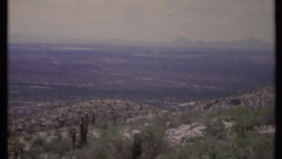1971 California 8mm landscape frame showing a cactus foreground, rocky desert slope, hazy valley floor, and faint mountain shapes.