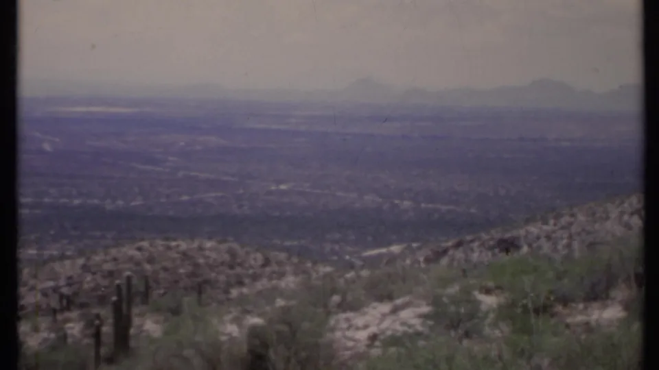 1971 California 8mm view from a rocky desert hillside with sparse cactus, a broad hazy valley, and mountains on the horizon.