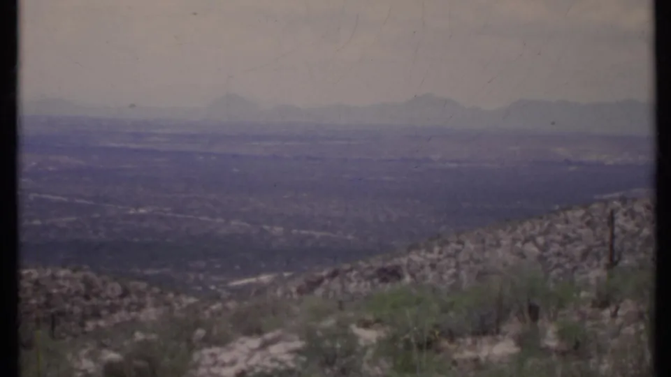 1971 California home-movie frame showing cactus and scrub on a rocky slope above a broad valley and hazy mountain horizon.