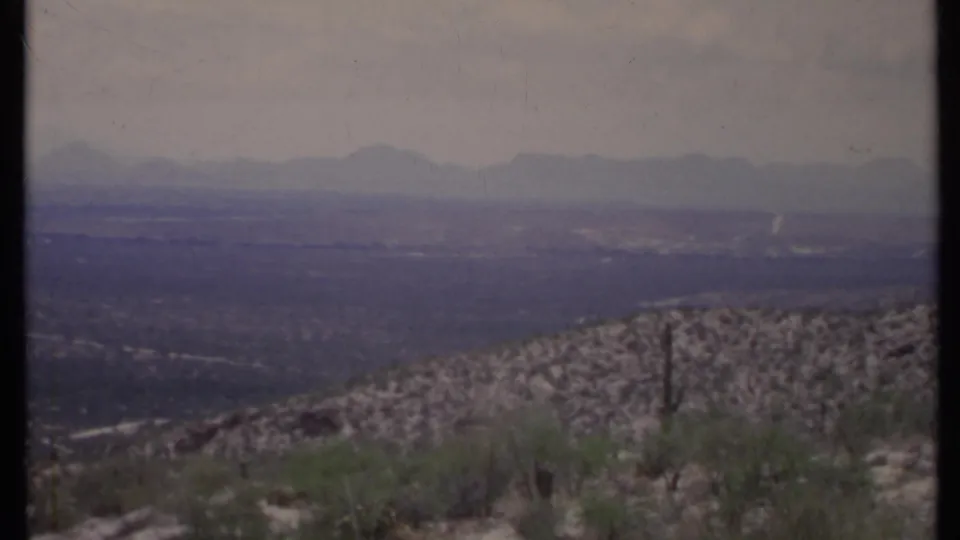 1971 California 8mm frame with a rocky foreground hill, scattered desert plants, valley floor, and soft mountain silhouettes.