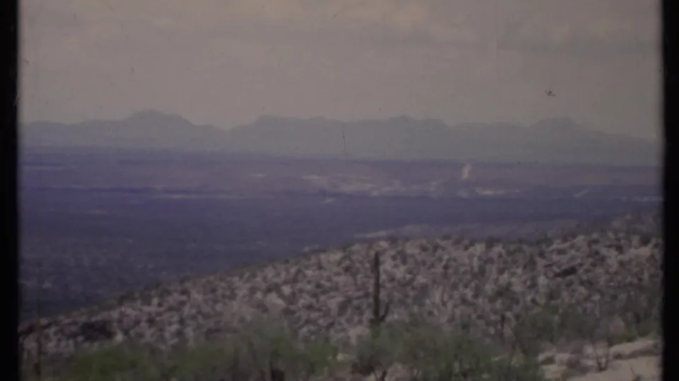 1971 California home-movie view across a rocky cactus-dotted hillside toward a hazy valley and distant mountains.