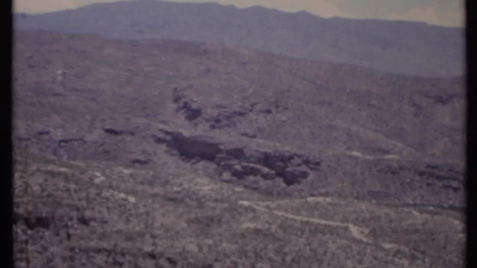 1971 California desert slope with a dark canyon-like formation, pale washes, sparse vegetation, and distant mountains.