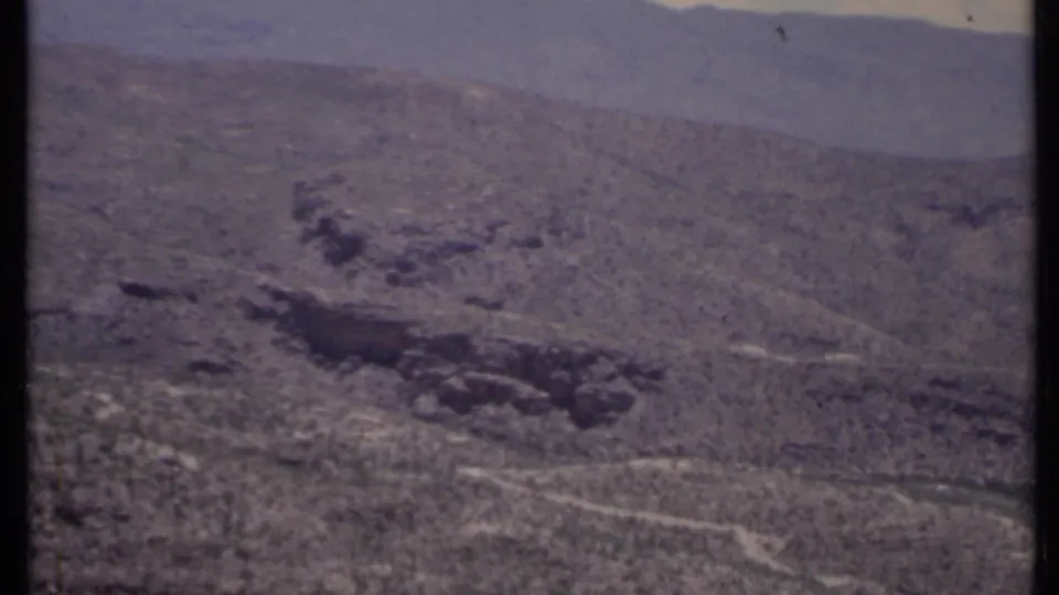 Vintage 8mm California desert view showing a dark rock formation, scrubby hillside, faint pale tracks or washes, and hazy mountains.