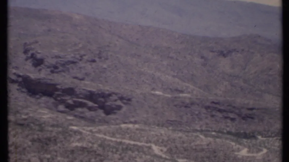 1971 California 8mm frame of an arid slope, dark canyon feature, winding pale wash, and distant hazy ridges.