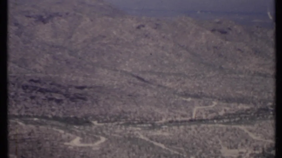 1971 California 8mm frame of an arid hillside landscape with brush, rocky textures, and pale dirt routes.