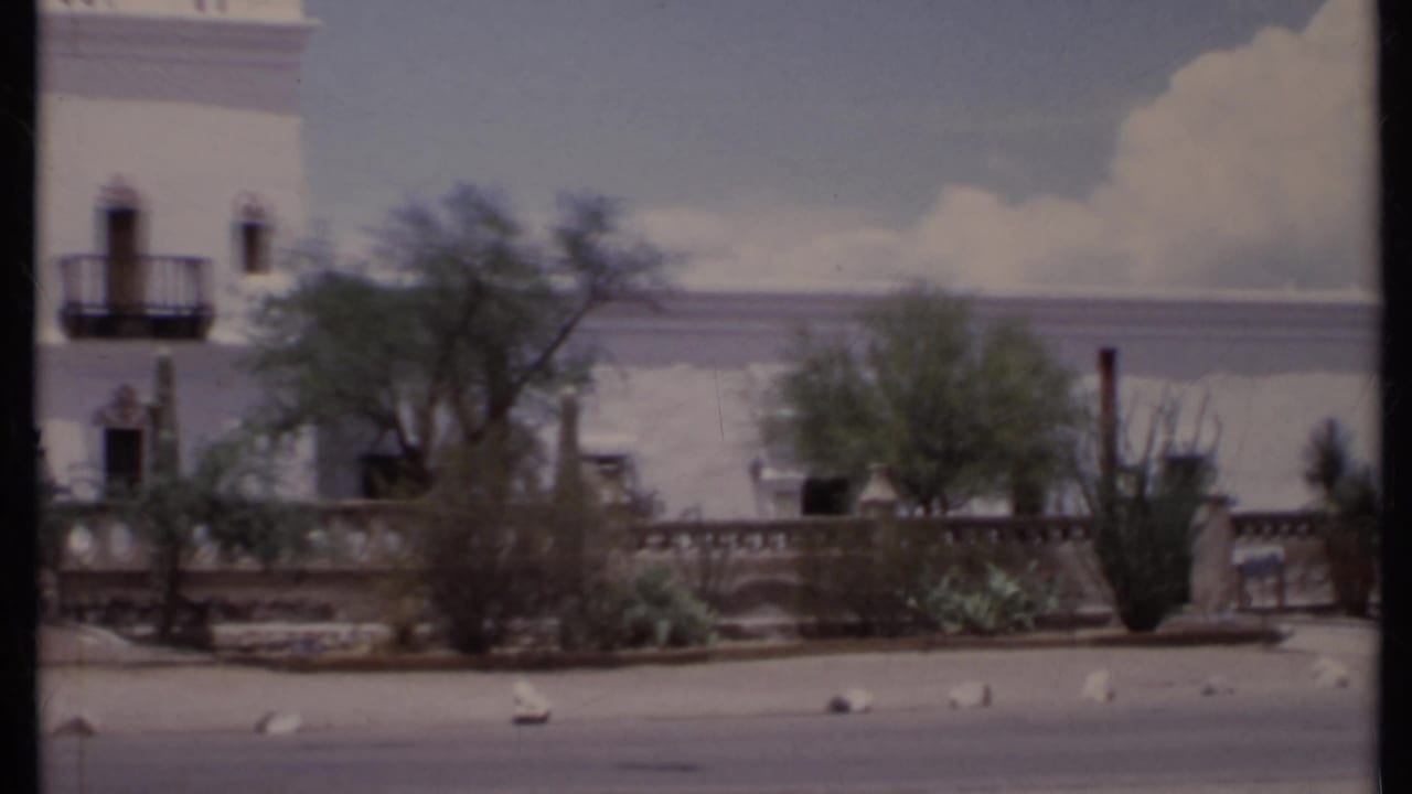 White stucco mansion facade with balconies, trees, cacti, low wall and street foreground.