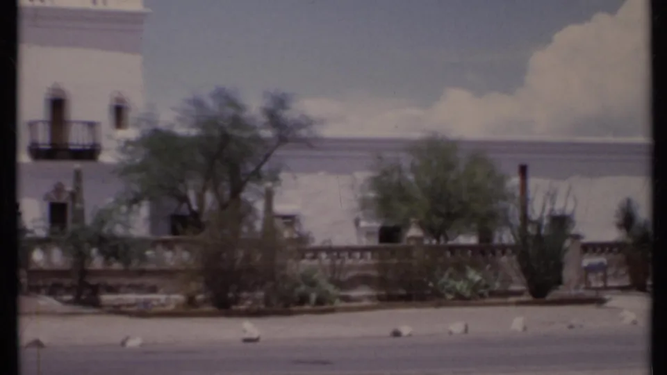 1971 California 8mm frame showing a white Spanish-style facade with small balconies, trees, cacti and a low wall by the street.