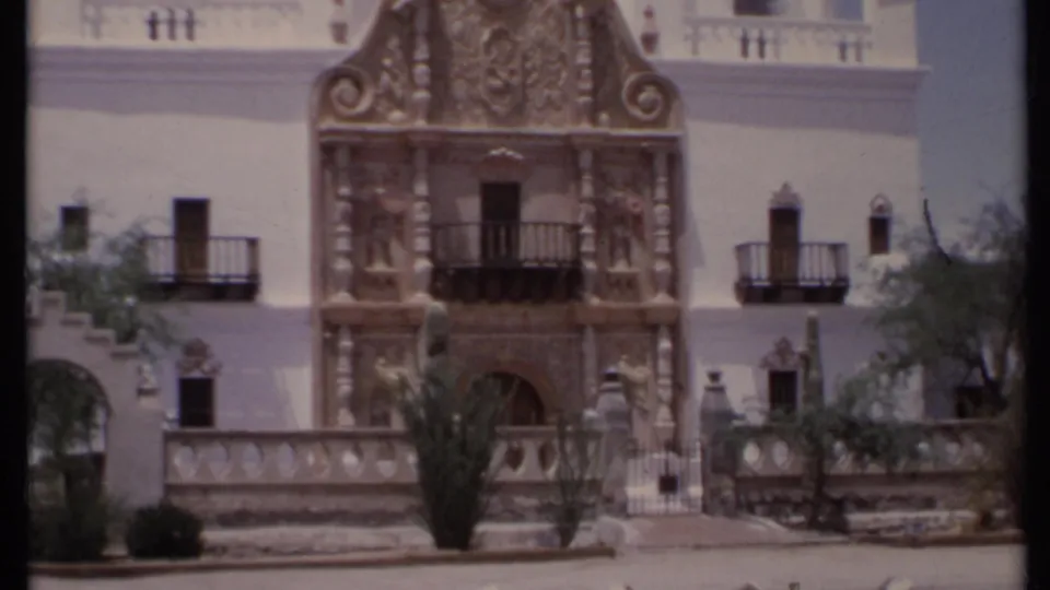 1971 California 8mm frame showing the ornate facade and surrounding white stucco architecture, with balconies, cacti and a low wall in front.