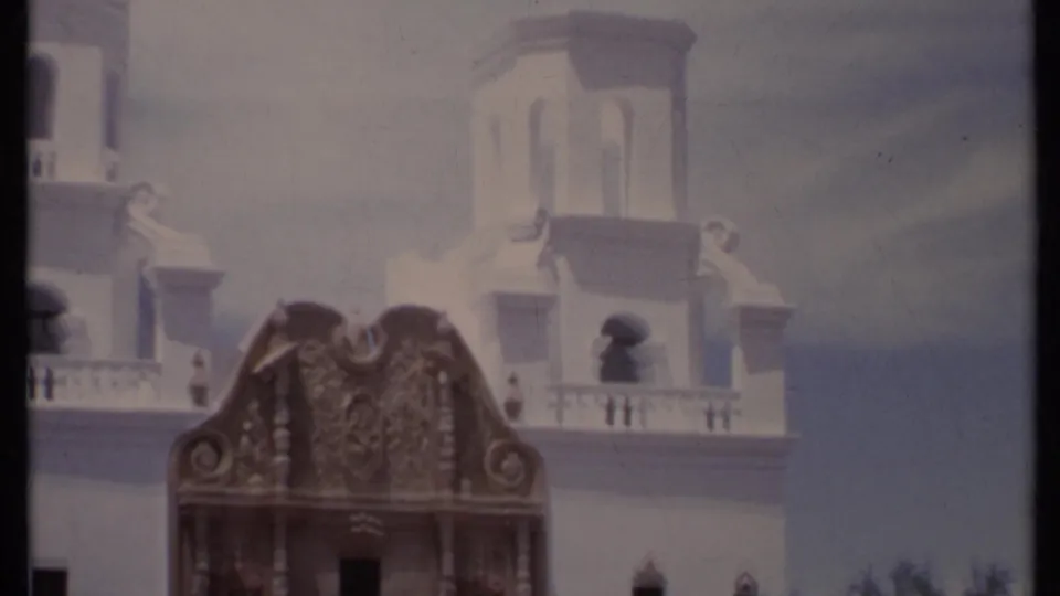 1971 California 8mm frame catching the upper white stucco structure and ornate facade top, with rooflines, balcony rail and hazy sky.