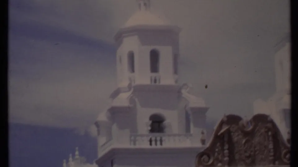 1971 California home-movie frame with a white towered facade, arched balcony area, and dark carved wooden architectural detail in the foreground.