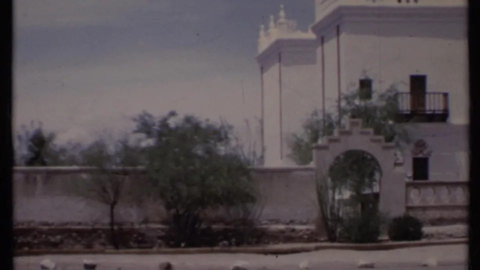 1971 California frame showing a white stucco building beside a low wall, with a balcony, arched opening, sparse trees, and the edge of a roadway.