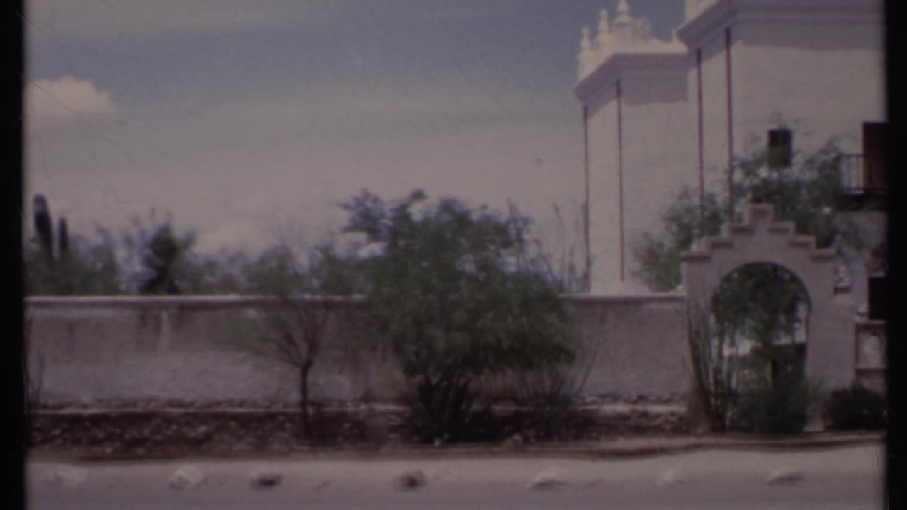 1971 California 8mm frame showing a white adobe-style exterior, arched gateway, wall, roadside pavement, and sparse desert planting.