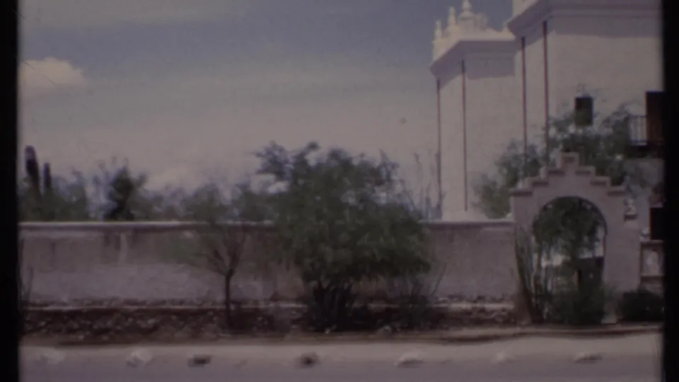 1971 California 8mm frame showing a white adobe-style exterior, arched gateway, wall, roadside pavement, and sparse desert planting.