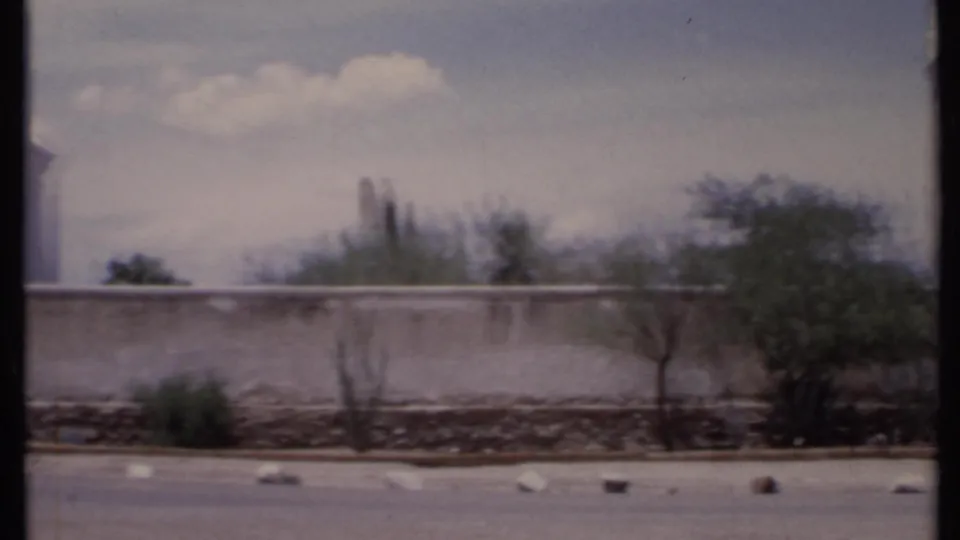 1971 California home-movie frame with a white wall, arched entry, tower-like structure, desert vegetation, and paved foreground.