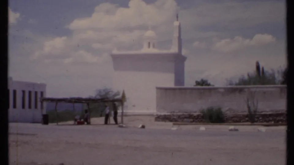 1971 California 8mm frame showing a white adobe-style structure, perimeter wall, shelter, and small distant people near the roadside.