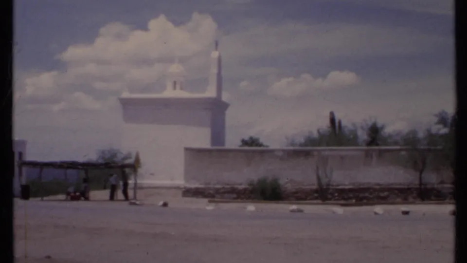 1971 California home-movie frame of a white adobe-style exterior, wall, sparse landscaping, shelter, and distant people at left.
