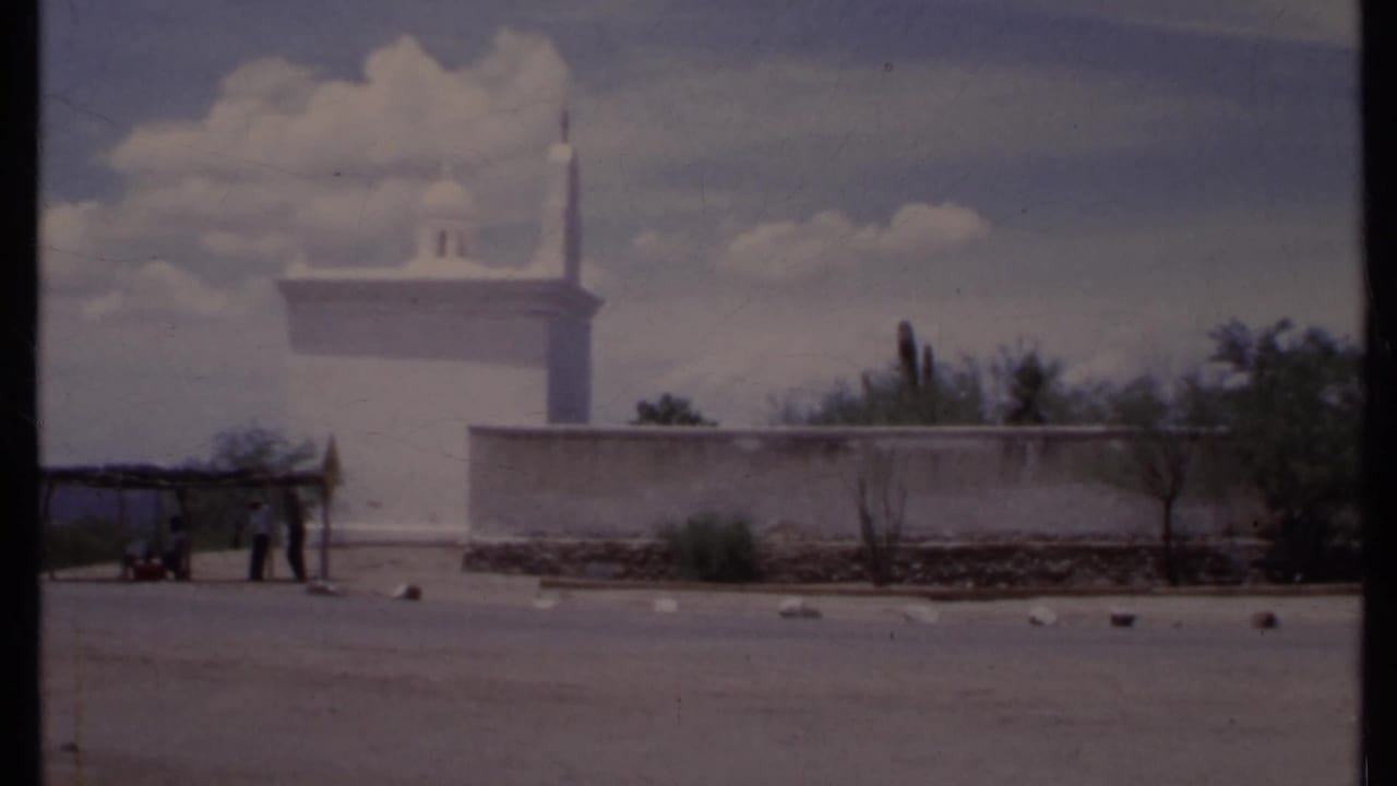 White adobe-style wall and tower across a paved lot, with a small shelter and people at left.