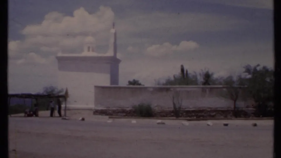 1971 California 8mm frame showing a white adobe-style exterior, minaret-like spire, shelter, distant people, and sunny parking area.