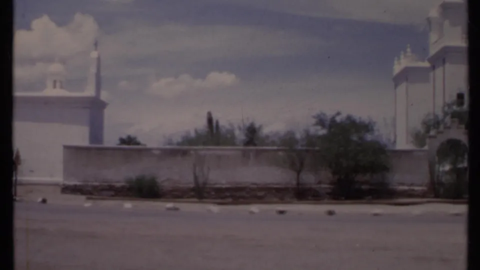 1971 California 8mm frame with a white wall, minaret-like spire, sparse trees, open parking area, and partial arched entrance.