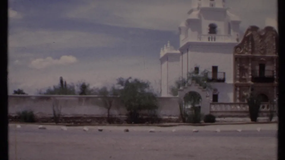 1971 California 8mm frame with a towered adobe-style exterior, arched gateway, desert shrubs, and sunlit parking area.